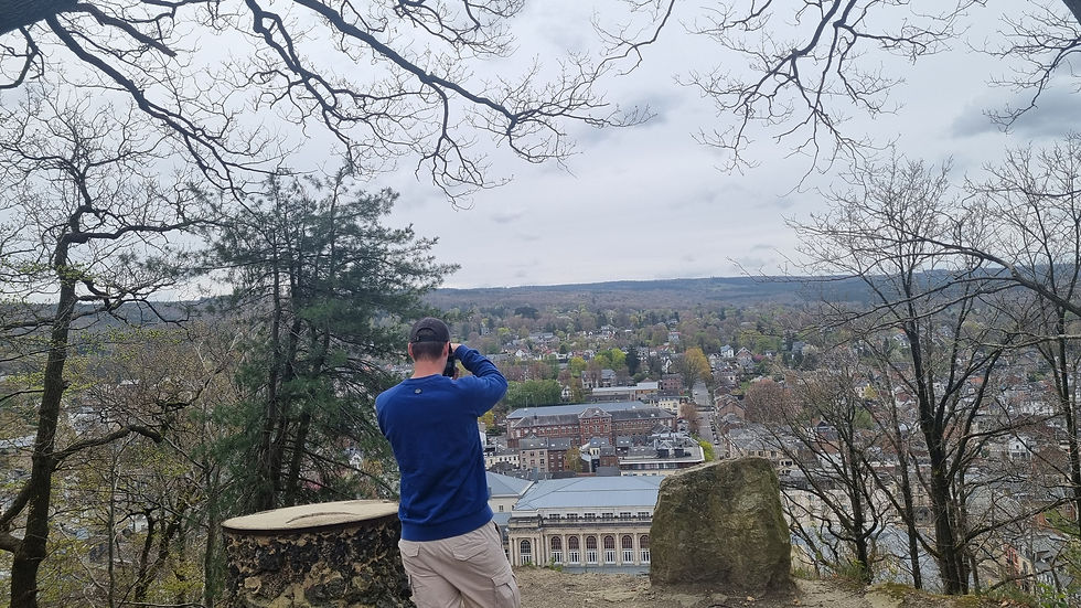 Man in blauwe trui maakt foto van stadspanorama. Hij staat op een heuvel, omringd door kale bomen en steen. Grijze lucht op achtergrond.