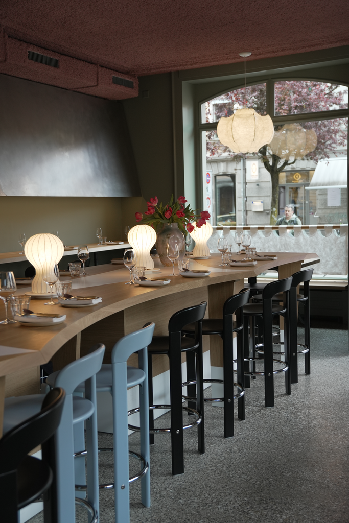 View of Capet Zurich’s stylish bar area with a curved wooden counter, modern high chairs, and softly glowing table lamps