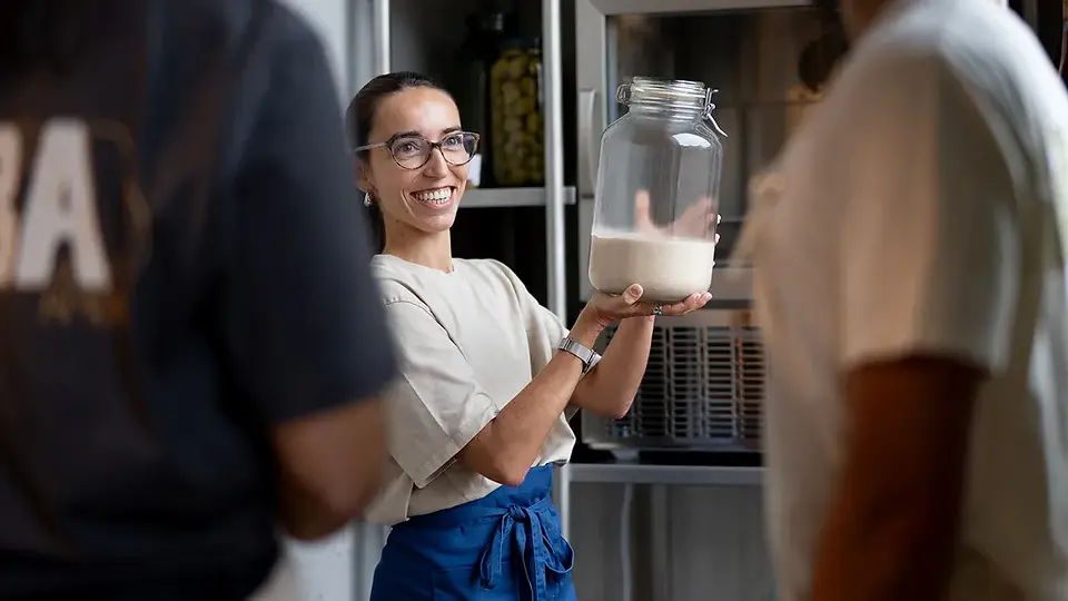 Smiling team member at ALBA Zurich holding a jar of sourdough starter, showcasing the behind-the-scenes passion — highlighted by BYCA Zurich.