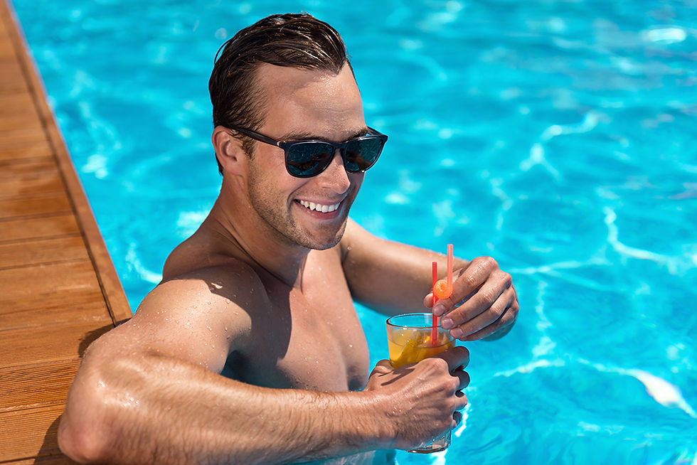 Man in sunglasses smiling while in a pool, holding a cocktail with a straw and garnish. Bright blue water, wooden pool edge. Relaxed mood.