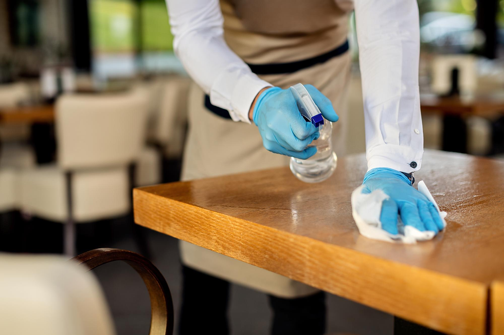 Medewerker in blauwe handschoenen ontsmet een houten restauranttafel met spray en doek.