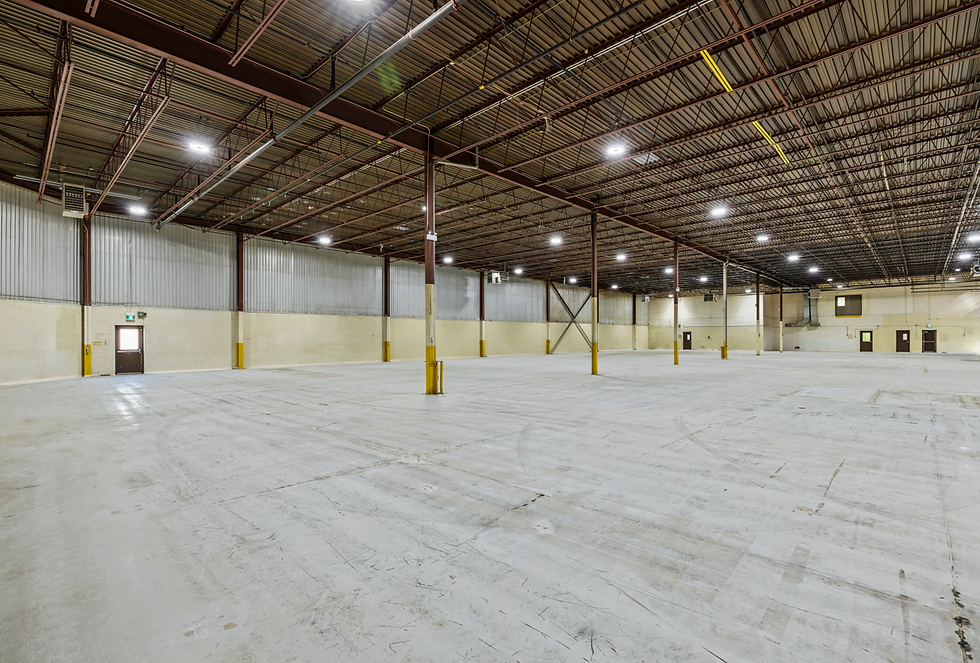 Empty warehouse interior with high ceilings, metal beams, and concrete floors. Bright lighting and yellow safety stripes on columns.