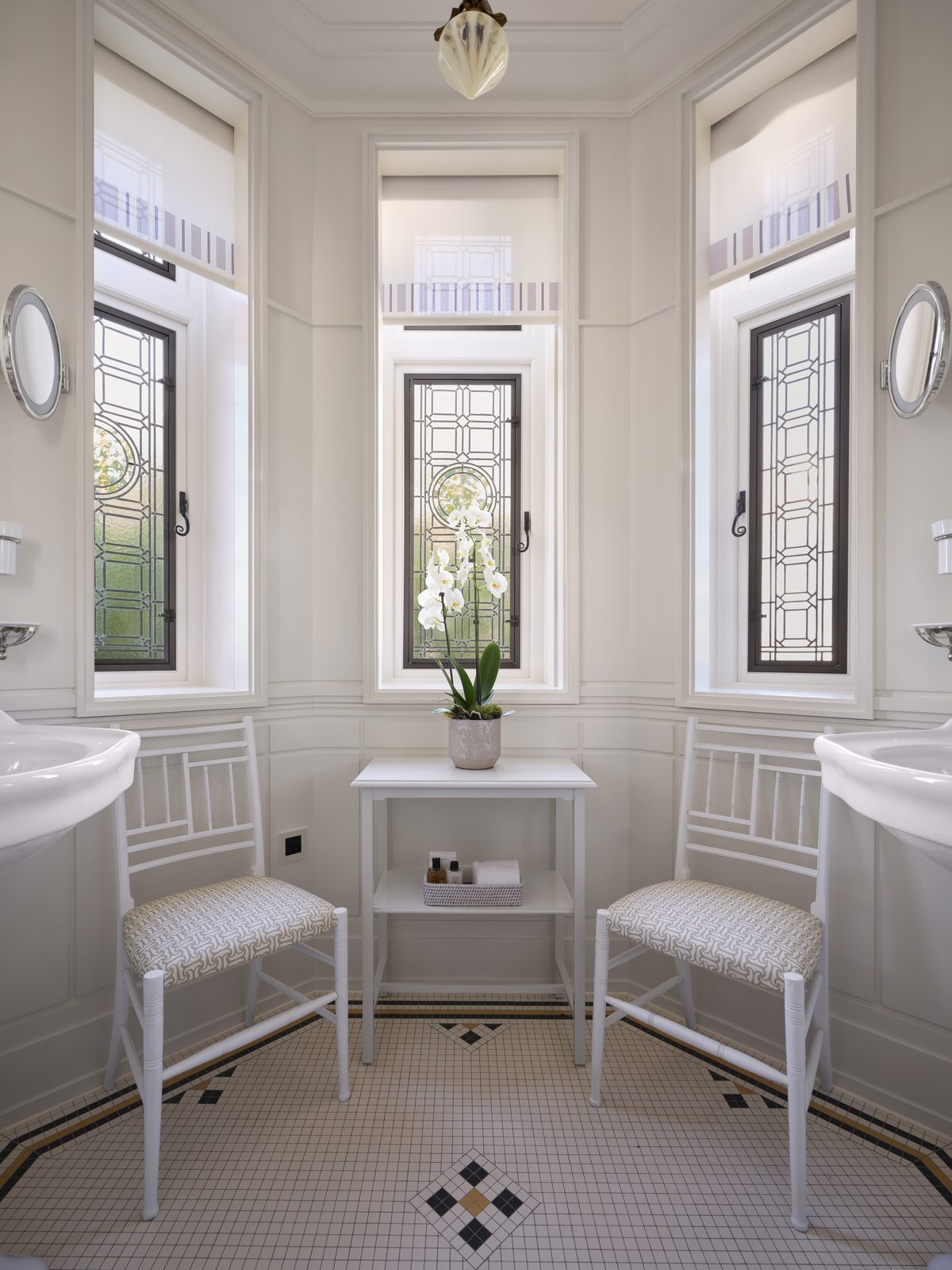 White bathroom in a bay with leaded-glass windows, a shell-glass ceiling light, twin basins and painted chairs