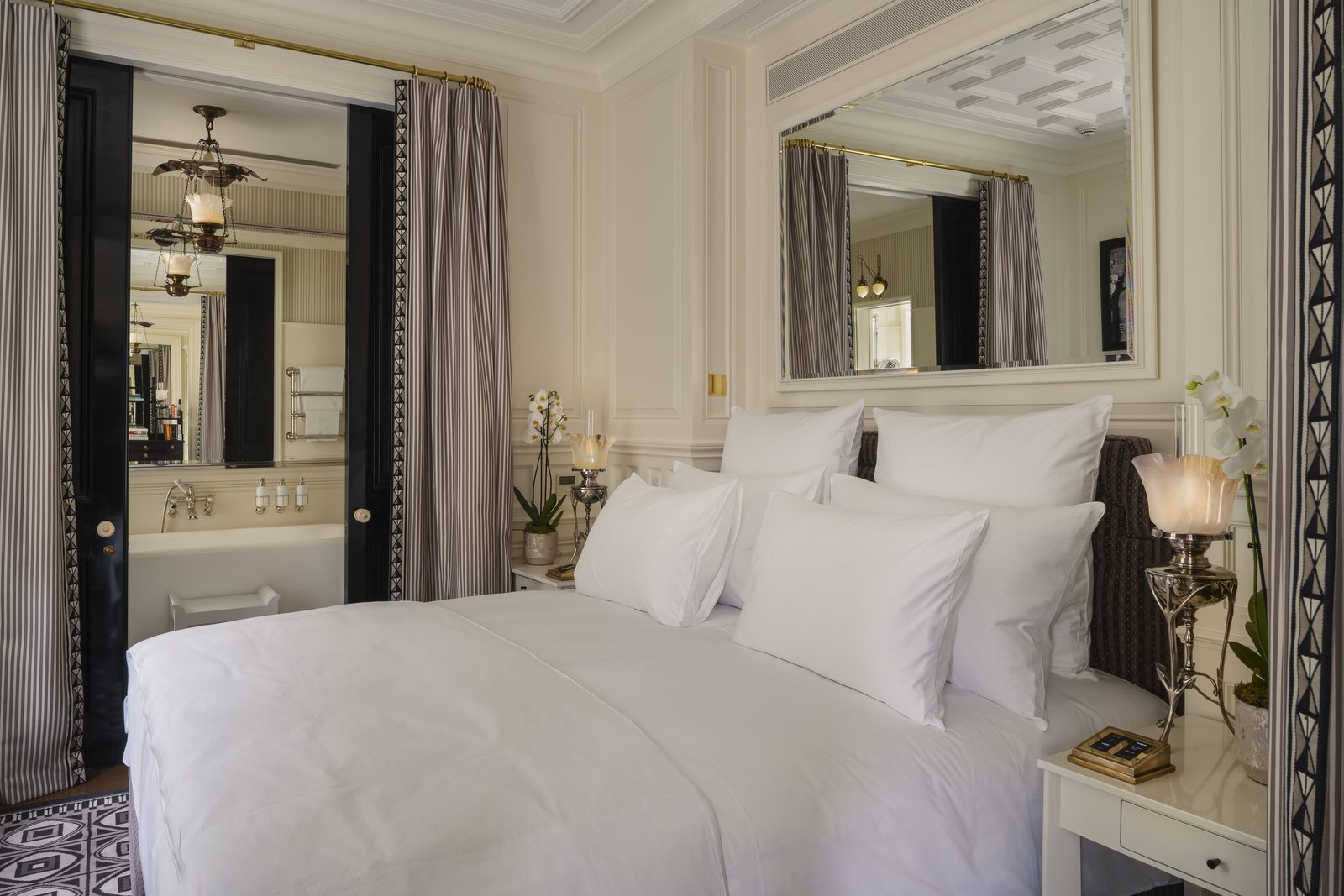 Panelled bedroom with striped curtains, a large mirror and glass-shaded table lamps, looking through to a bathroom with brass and glass lanterns