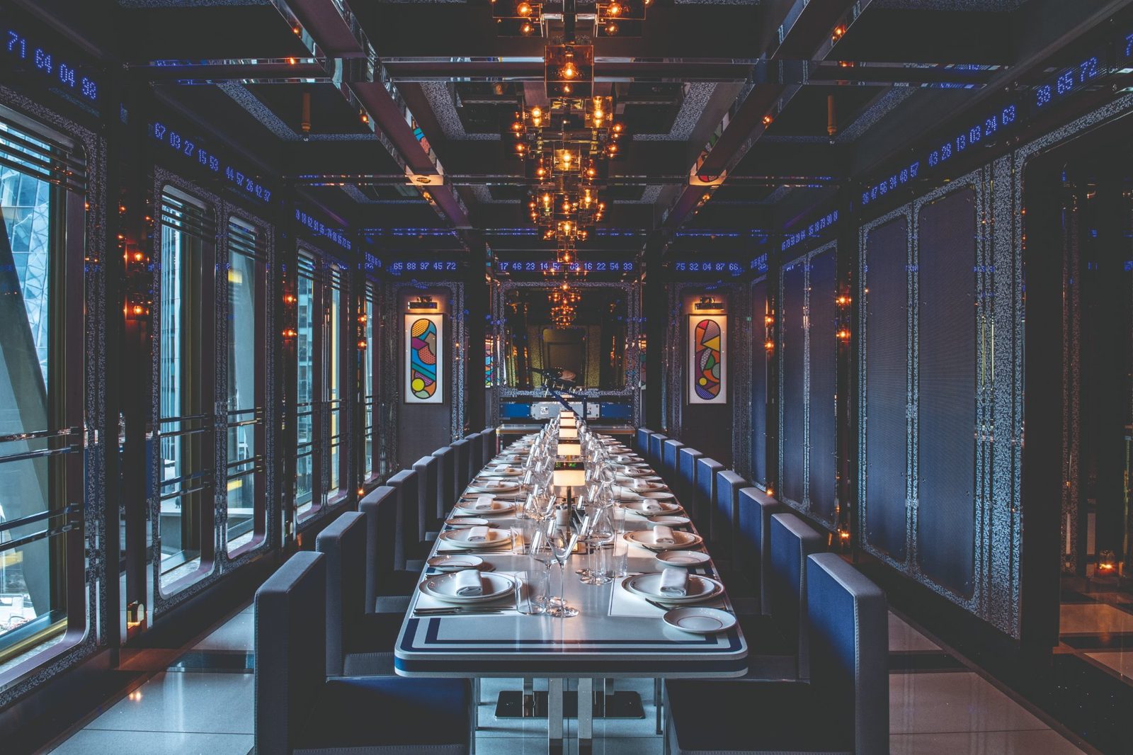 Private dining room with a long table beneath a run of cube-shaped glass and metal pendants, with lit wall panels and city views