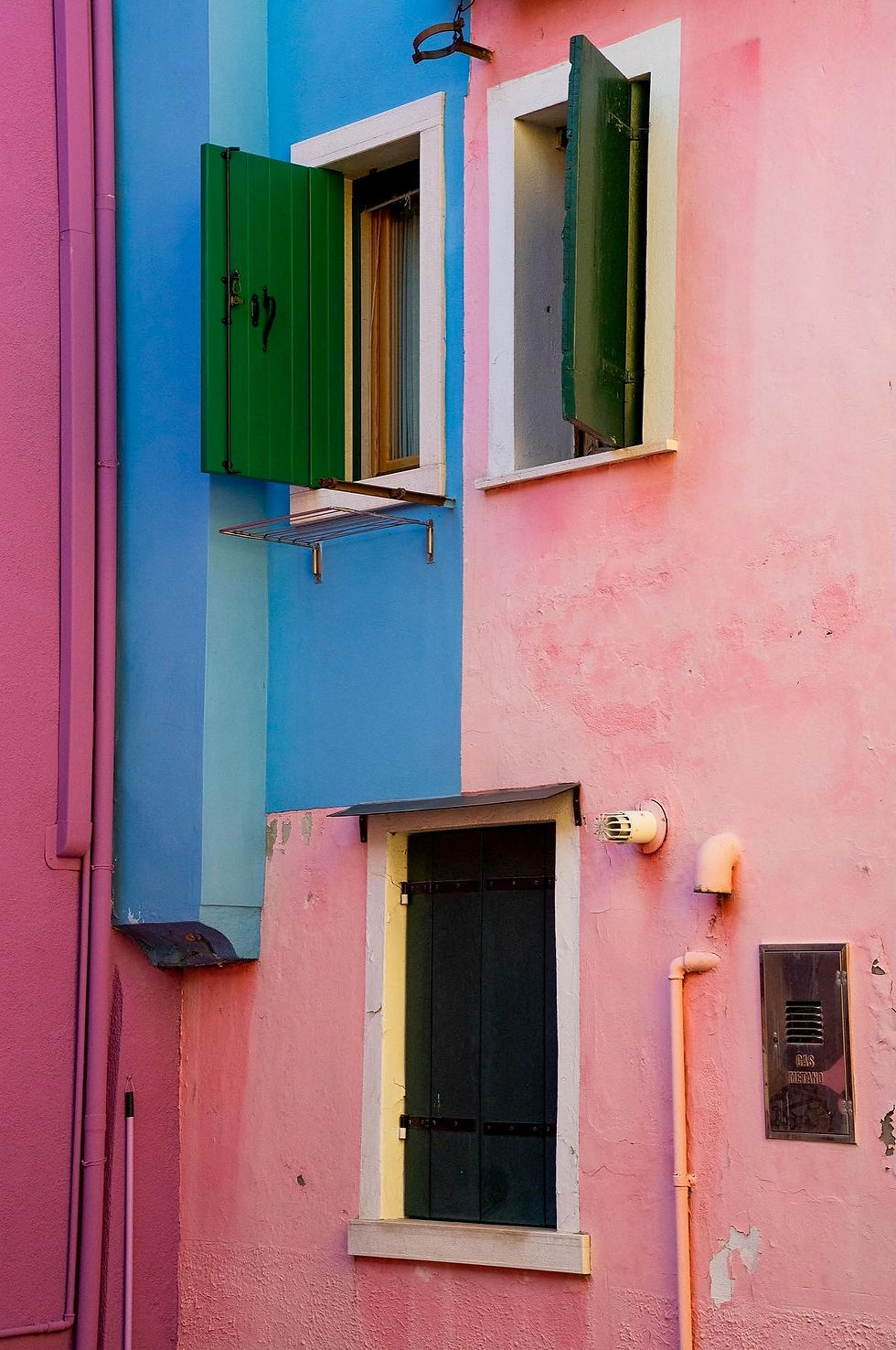 Photographie d’une façade colorée de Burano avec murs bleu et rose, volets verts et textures patinées
