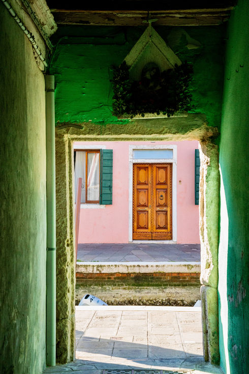 Photographie d’une façade rose à Burano vue à travers un passage aux murs verts patinés
