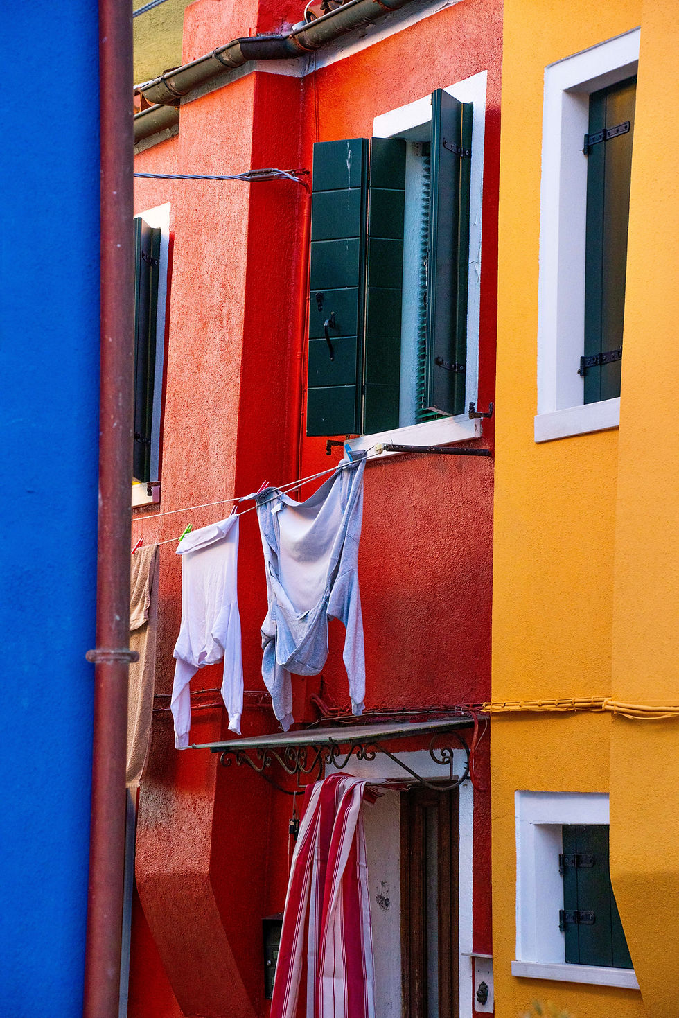 Photographie d’art d’une façade colorée à Burano avec linge suspendu, volets verts et contrastes entre bleu, rouge et jaune,