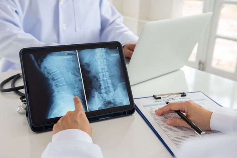 Doctor examines spinal X-rays on a tablet while another takes notes on a clipboard. Neutral setting, white coats, medical consultation.