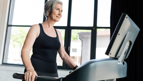 Smiling older woman in black workout clothes uses a treadmill in a bright gym by large windows.