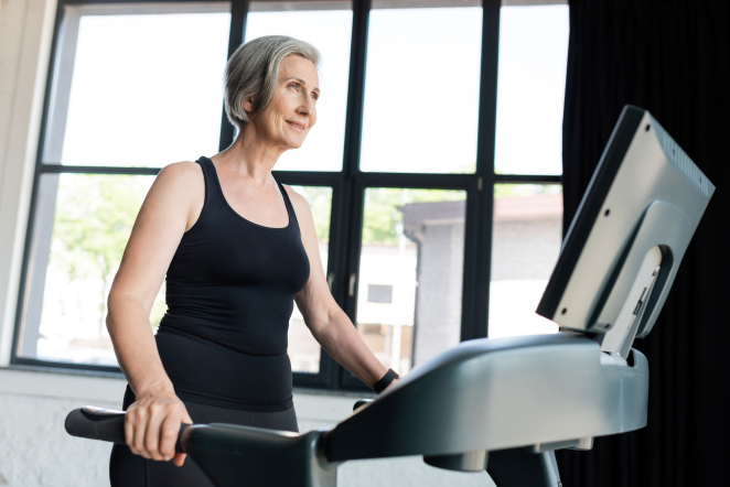 Smiling older woman in black workout clothes uses a treadmill in a bright gym by large windows.