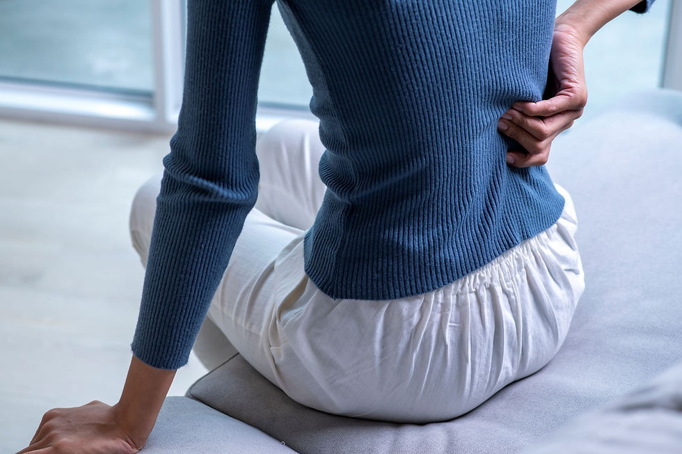 Person in blue sweater and white pants sits on a bed, clutching lower back in a bright room, suggesting back pain.