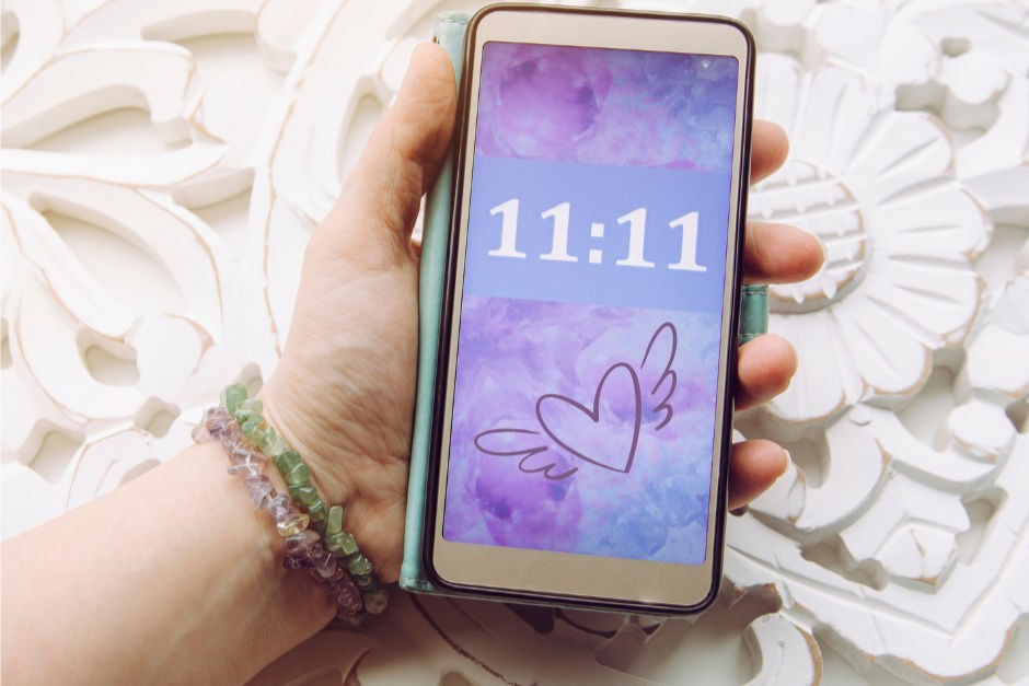 Hand holding a phone with a purple screen showing "11:11" and a heart with wings. Background has white floral patterns. Wrist has crystal bracelets.