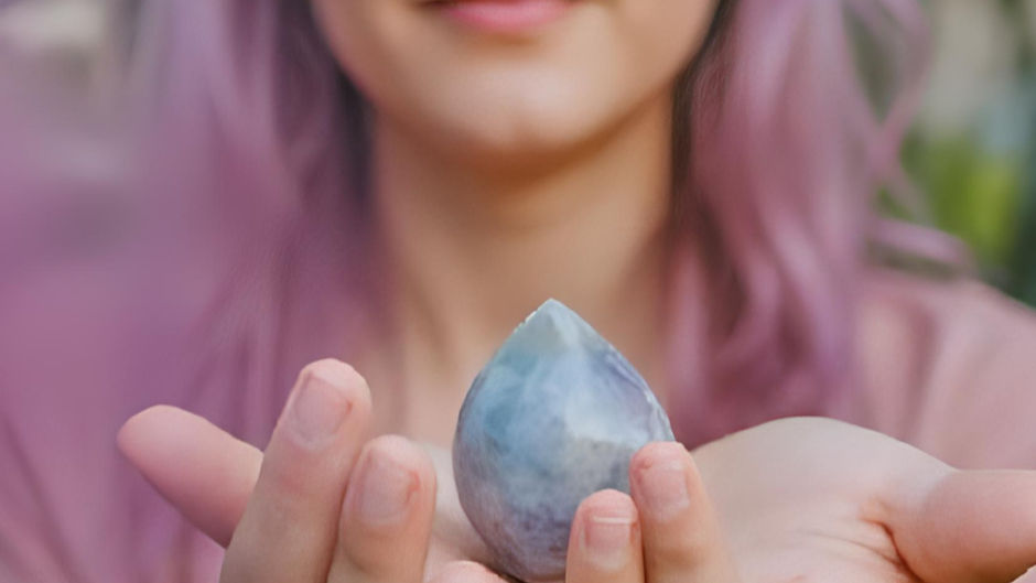 Person with pink hair holds a smooth, blue stone in outstretched hands. Background is blurred, creating a tranquil mood.