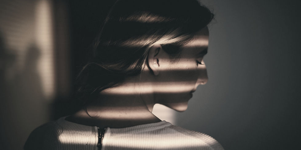 Woman in white top, standing in dim light with blinds casting striped shadows across her face, creating a contemplative mood.