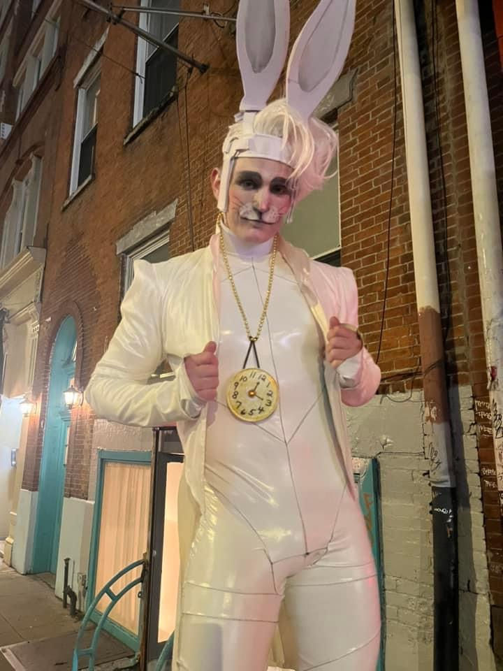Person in White Rabbit costume holding jacket, wearing necklace, near brick wall.