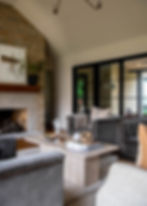 Cozy living room with gray sofas, a stone fireplace, and a cream marble coffee table. Decor includes books, a candle, and potted plants.