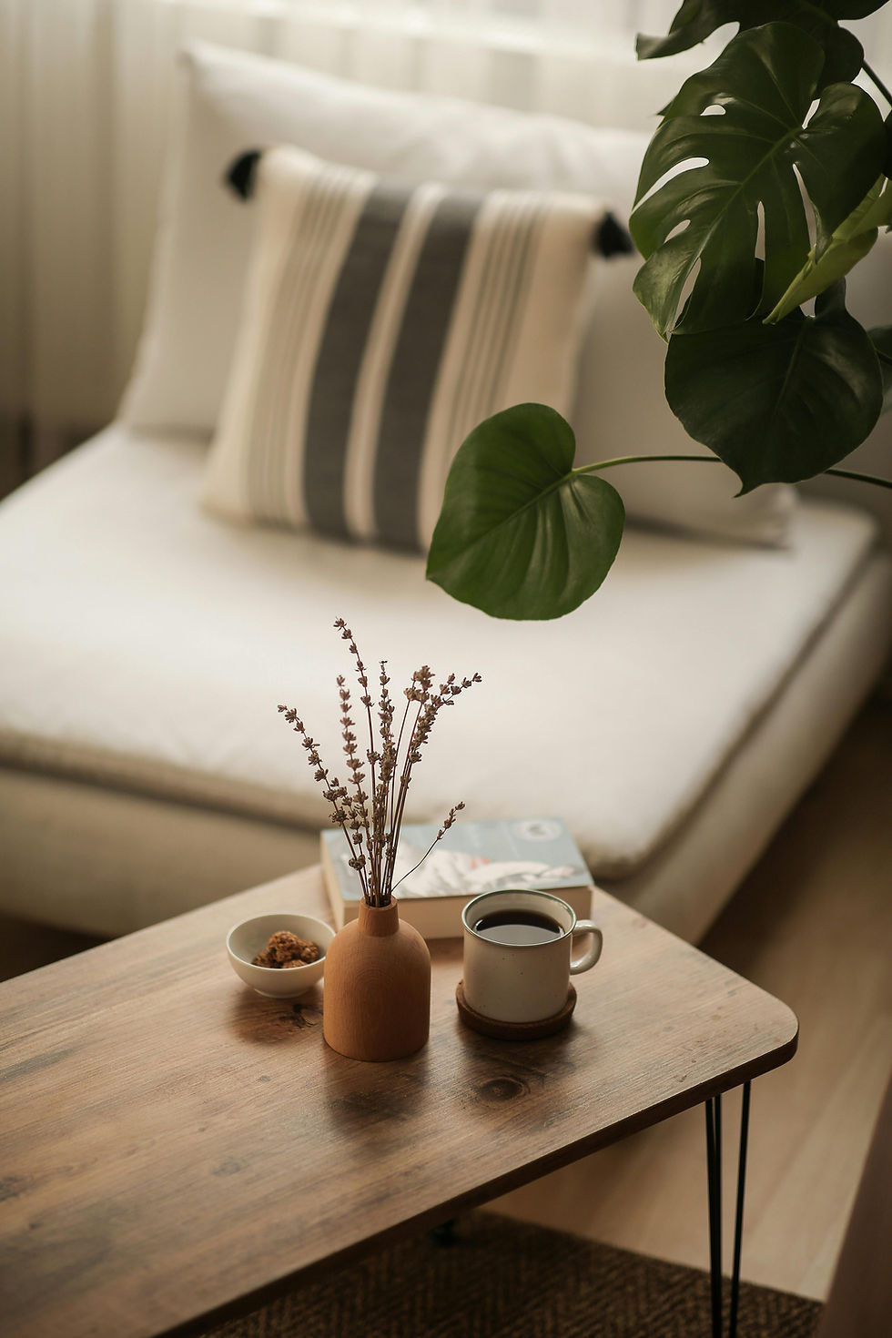 Een houten salontafel met een vaas bloemen, koffie, en snacks. Achtergrond is een bank met gestreept kussen en een grote groene plant. Rustige sfeer.
