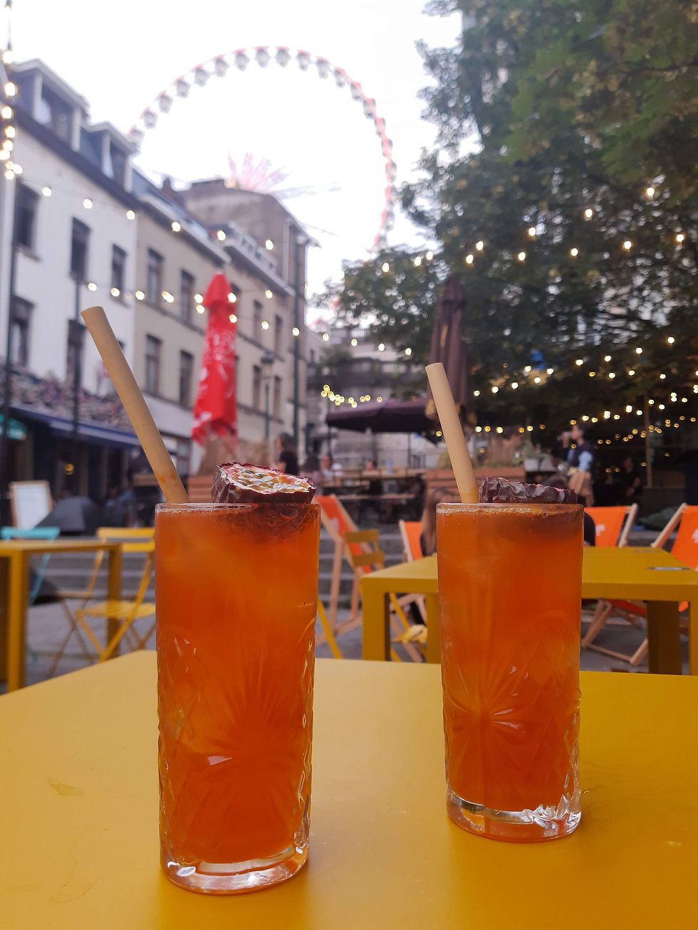 Twee oranje cocktails op een gele tafel met een reuzenrad op de achtergrond. Sfeervolle verlichting boven terras in stedelijke omgeving.