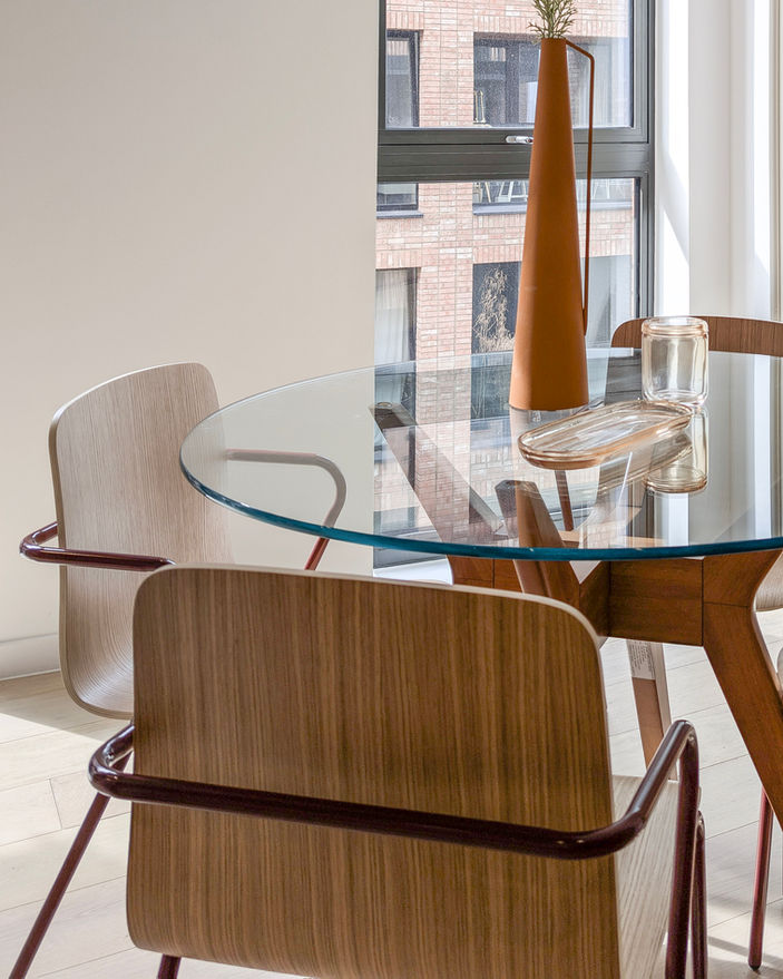 Contemporary dining space with round glass table, wood and metal chairs and natural light flooding through black-framed windows.