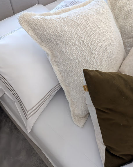 Detailed view of crisp white bed linen layered with neutral and olive-toned cushions against a soft upholstered headboard.