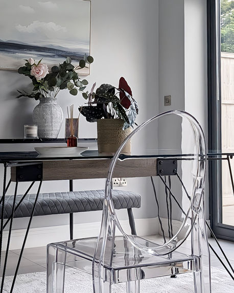 Dining corner with transparent acrylic chair, wooden table, and floral arrangement positioned near large windows for natural light emphasis.