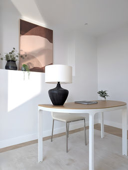 Home office setup with oval desk, modern table lamp, and warm-toned abstract artwork in light-filled apartment.