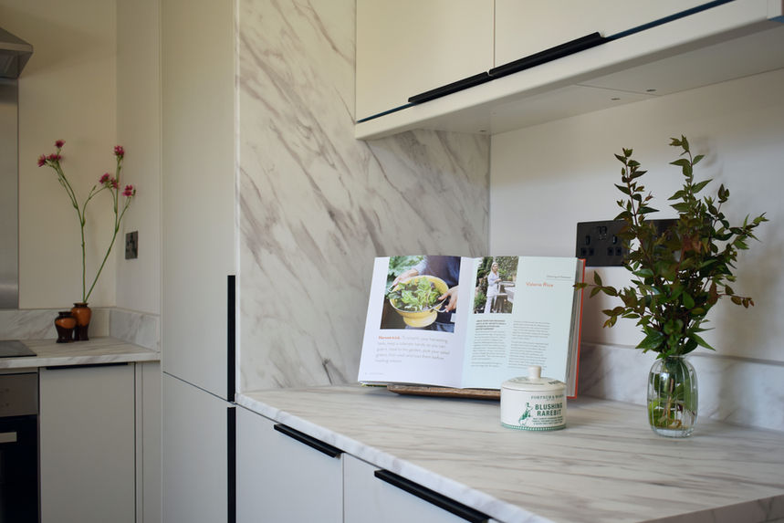 Contemporary kitchen with marble backsplash, handleless cabinetry, open cookbook display, and fresh greenery arranged along the countertop for lifestyle-focused staging.