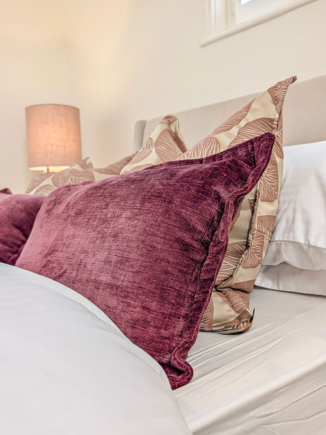 Close-up of upholstered headboard with layered velvet cushions in deep plum and soft neutrals, styled for a Victorian-inspired interior.