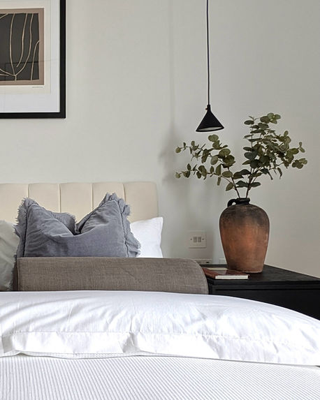 Bedroom vignette with upholstered headboard, layered neutral bedding, and bedside plant detail.