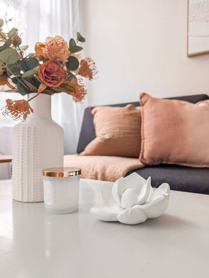 Close-up of a marble coffee table styled with a white ceramic vase, candle, and warm-toned floral arrangement in front of a charcoal sofa.