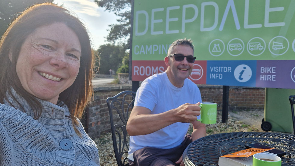 Smiling couple at an outdoor café by a Deepdale Camping sign, man raising a green mug, sunny relaxed mood.