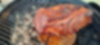 Grilled meat on a charcoal BBQ with a rich, brown crust. Gray ash and coals visible through the grill. Warm and rustic setting.