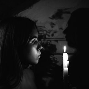 « Photographie d'une femme de profil, le visage doucement éclairé par la lueur d'une bougie dans l'obscurité. Elle évoque la figure de la sorcière ou la gardienne des secrets.