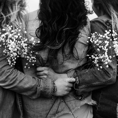 Trois femmes de dos en chemises en jean se tenant par la taille, portant des bouquets de fleurs blanches. Ambiance amicale et sereine pour illustrer la sororité du Women's Prize for fiction