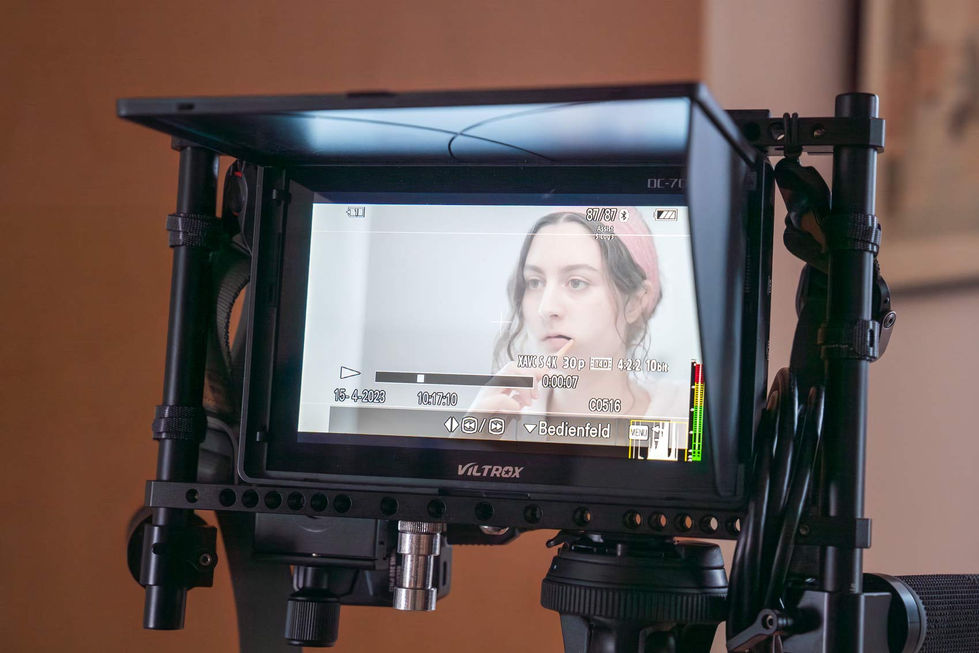 Eine Nahaufnahme des Bildschirms einer Filmkamera zeigt eine junge Frau mit einem rosa Stirnband, die vor einem Spiegel steht. Der Bildschirm zeigt detaillierte Aufnahmeinformationen und ein klares Bild des Motivs. Die Kamera ist professionell ausgestattet und auf ein Stativ montiert.