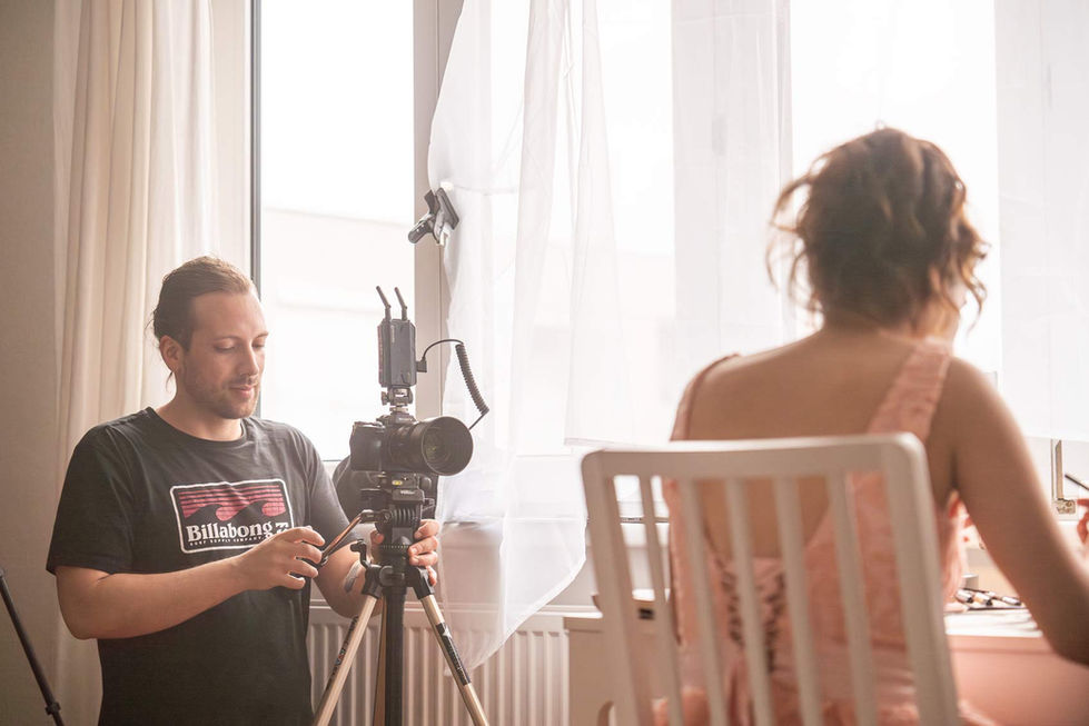Ein Mann mit einem schwarzen Billabong-T-Shirt steht hinter einer Kamera auf einem Stativ und richtet diese aus. Er befindet sich in einem Raum mit großen Fenstern und weißen Vorhängen. Im Vordergrund sitzt eine Frau mit hochgesteckten Haaren und einem rosa Spitzenkleid auf einem Stuhl und ist teilweise im Bild zu sehen. Die Szene zeigt ein Film- Fotoshooting mit einem professionellen Kamerasetup.