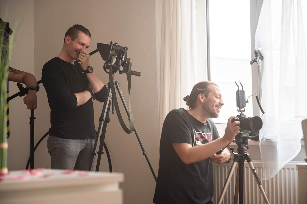 Zwei Männer stehen in einem Raum mit Kameraequipment. Der Mann links, in einem schwarzen Shirt und grauen Jeans, lacht und hält eine Hand vor sein Gesicht. Der Mann rechts, in einem schwarzen Billabong-T-Shirt, kniet und bedient eine Kamera auf einem Stativ, während er ebenfalls lächelt. Im Hintergrund sind weiße Vorhänge und ein Fenster zu sehen. Die Szene zeigt eine entspannte und fröhliche Atmosphäre während eines Film- oder Fotoshootings.