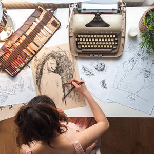 Ein Mädchen sitzt an einem Schreibtisch und zeichnet auf braunem Papier eine Skizze einer Figur. Auf dem Schreibtisch befinden sich verschiedene Zeichnungen, ein Schreibmaschinen-Modell, eine Pflanzen-Dekoration, eine Kerze, eine Auswahl an Zeichenstiften und Skizzenblöcken. Die Atmosphäre wirkt kreativ und künstlerisch.