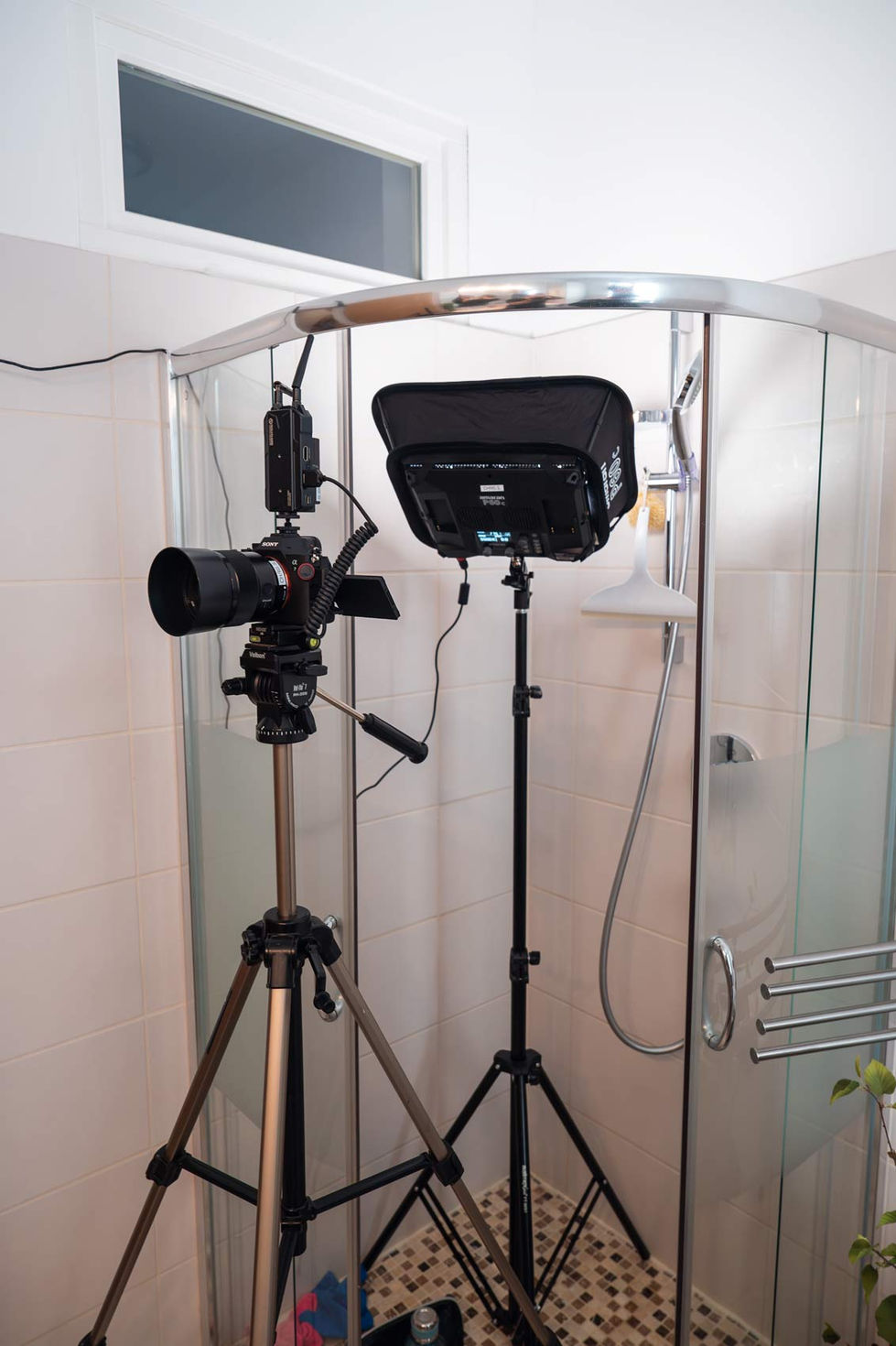 Eine Duschkabine, die für ein Film- oder Fotoshooting eingerichtet ist. In der Duschkabine steht ein Stativ mit einer montierten Kamera und ein weiteres Stativ mit einer Beleuchtungseinheit, die mit einem Softbox-Diffusor ausgestattet ist. Kabel verlaufen entlang der Wände, um die Geräte mit Strom zu versorgen. Die Szene zeigt die technische Vorbereitung und den Aufbau für eine Aufnahme in einem Badezimmer.