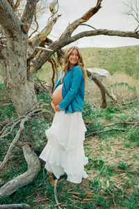 Pregnant woman in white dress and denim shirt standing among desert trees during maternity session at Mission Creek Preserve near Joshua Tree