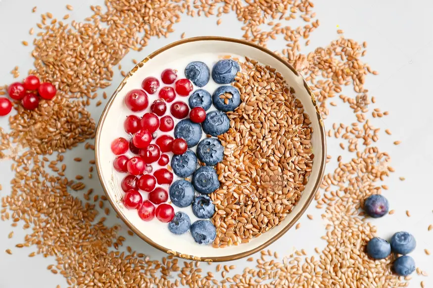 A bowl of creamy yogurt topped with flax seeds, fresh blueberries, and red currants, surrounded by scattered flax seeds on a light background.