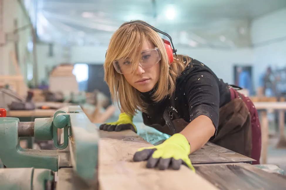 Woman in safety gear sands wood in a workshop, focused and determined. She's wearing earmuffs, goggles, and gloves. Background is blurred.