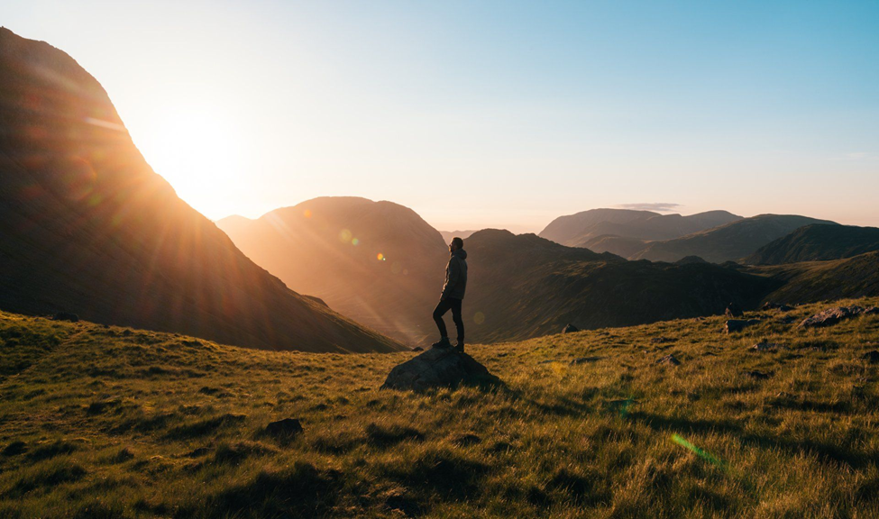 Silhouette of a person stands on a rock, overlooking a sunlit valley with grassy hills and mountains. The sun sets, casting a warm glow.