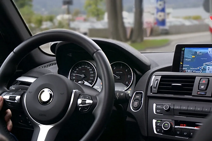 Car interior showing a hand on the steering wheel, dashboard with speedometer, and GPS screen displaying a map. Trees visible outside.