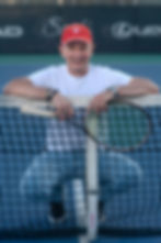 A man in a white shirt and red cap kneeling behind a tennis net while holding a tennis racket on an outdoor court.