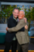 Two men warmly hugging outdoors with string lights glowing in the background.