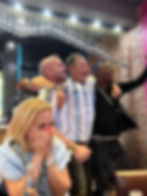 A group of people cheering energetically in a restaurant while a woman in the foreground covers her mouth in emotion.