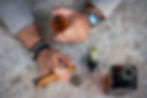 Close-up of a person’s hands holding a cigar and a glass of whiskey on a textured tabletop.