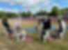 5 women on white plastic chairs with their arms raised, face another woman on a chair who demonstrates, they are outside on grass